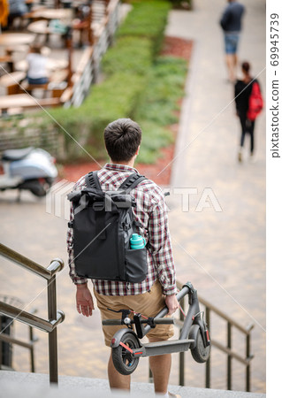 Man walking with folded electric scooter up stairs in city near modern building. Ecological technological lifestyle. E-Mobility. Person Carrying Electric Scooter Folded Position. Eco urban transport 69945739