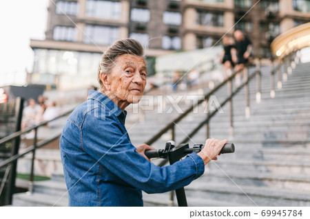 Active old woman riding electric scooter. Retired lady uses environmentally friendly city vehicle. Granny very old with gray hair, active and progressive. Modern senior woman use technology e scooter Active old woman riding electric scooter. Retired lady uses environmentally friendly city vehicle. Granny very old with gray hair, active and progressive. Modern senior woman use technology e scooter 69945784