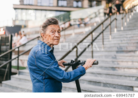 90 year old woman with gray hair, wrinkles, progressive and active uses modern electric transport scooter. Lady pensioner use eco friendly city vehicle. Senior female using urban personal transport 69945787