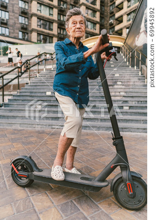 Cheerful elderly woman on a scooter. Grandma rides a scooter. Grandmother using urban personal transporter. Active retirement concept. Healthy cheerful senior retired lady. Ecological transport Cheerful elderly woman on a scooter. Grandma rides a scooter. Grandmother using urban personal transporter. Active retirement concept. Healthy cheerful senior retired lady. Ecological transport 69945892