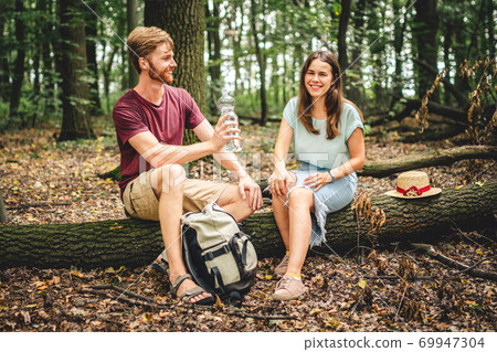 Couple relaxing sitting on a log and drinking water. The family is enjoying a romantic weekend in nature. A pleasant walk or a romantic date in the background of nature. Picnic hike in the forest 69947304