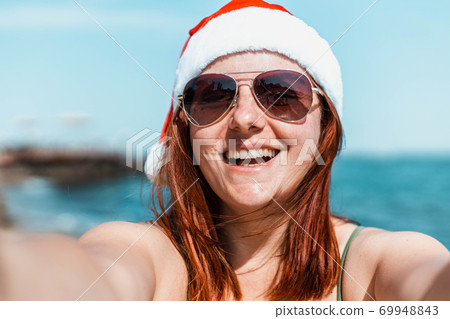 Beautiful happy girl in santa hat and swimsuit makes selfie and enjoying winter holidays getaway on tropical beach. Beautiful happy girl in santa hat and swimsuit makes selfie and enjoying winter holidays getaway on tropical beach. 69948843