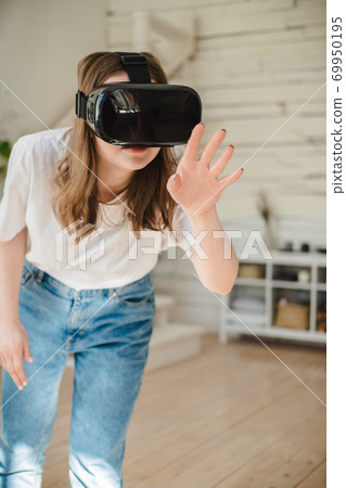 Young woman in jeans and white T-shirt wearing virtual reality helmet plays game, works, watches video in room next to bed. Concept of modern technology, VR, augmented virtual games, entertainment. 69950195