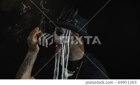 Scary guy in carnival costume of Halloween skeleton smoking cigar, making faces, showing tongue 69951263