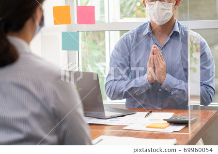 Asian business team colleagues wearing mask greeting by looking through the plastic protective wall, concept of social distancing to prevent the spread of coronaviru COVID-19. 69960054
