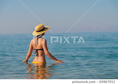 An attractive slender young Caucasian woman in a swimsuit and a straw hat stands waist-deep in sea water in the morning when there is haze over the sea An attractive slender young Caucasian woman in a swimsuit and a straw hat stands waist-deep in sea water in the morning when there is haze over the sea 69967052
