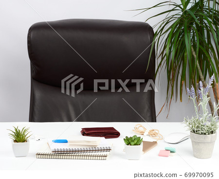 an empty brown leather armchair near a white table with a stack of notebooks, a purse and flowerpots with flowers an empty brown leather armchair near a white table with a stack of notebooks, a purse and flowerpots with flowers 69970095