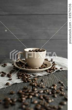 Coffee cup filled of fresh arabica or robusta coffee beans with scattered coffee beans on a linen textile and wood table. Coffee cup filled of fresh arabica or robusta coffee beans with scattered coffee beans on a linen textile and wood table. 69974436