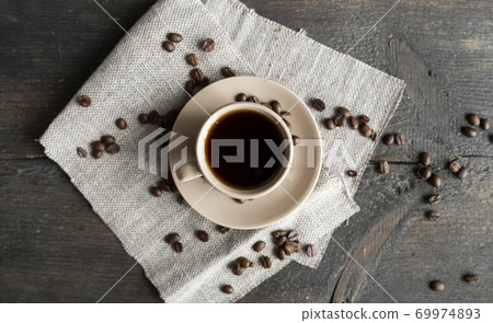 Coffee cup with scattered coffee beans on a wooden table background. Mug of black coffee on a linen textile. Coffee cup with scattered coffee beans on a wooden table background. Mug of black coffee on a linen textile. 69974893