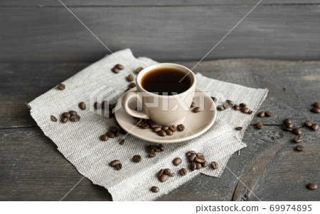 Coffee cup with scattered coffee beans on a wooden table background. Mug of black coffee on a linen textile. Coffee cup with scattered coffee beans on a wooden table background. Mug of black coffee on a linen textile. 69974895