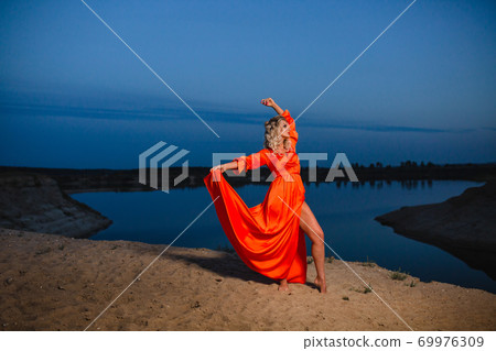 Young woman in bright orange dress posing at the background of blue sky Young woman in bright orange dress posing at the background of blue sky 69976309