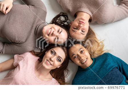Four happy diverse ethnicity pretty young girls women friends lying in circle look at camera together, sleepover party and multiethnic friendship concept, top view portrait 69976771