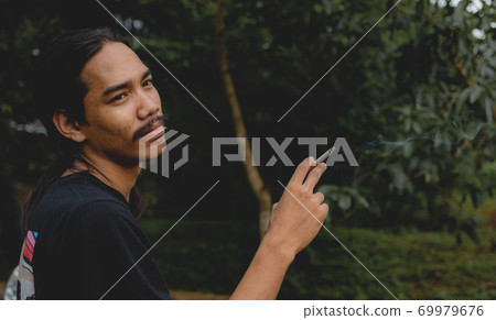 A long-haired Asian man with a beard holding a cigarette standing outside A long-haired Asian man with a beard holding a cigarette standing outside 69979676