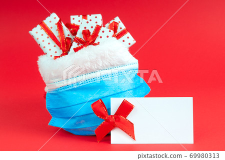 Christmas sack of Santa Claus with gifts in a protective face mask on a red background with note mock up. The concept of holidays during the coronavirus. Christmas sack of Santa Claus with gifts in a protective face mask on a red background with note mock up. The concept of holidays during the coronavirus. 69980313