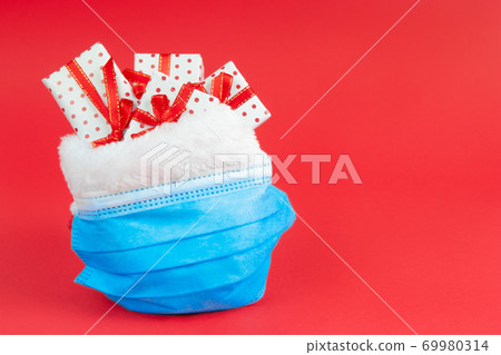 Christmas sack of Santa Claus with gifts in a protective face mask on a red background with copy space. The concept of holidays during the coronavirus. Christmas sack of Santa Claus with gifts in a protective face mask on a red background with copy space. The concept of holidays during the coronavirus. 69980314