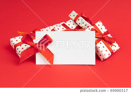 White gifts packed in wrapping paper pot with festive ribbon on red background with note mock up. White gifts packed in wrapping paper pot with festive ribbon on red background with note mock up. 69980317