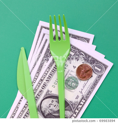 Disposable plastic cutlery green. Plastic fork and knife lie on a small amount of US dollars Disposable plastic cutlery green. Plastic fork and knife lie on a small amount of US dollars 69983894