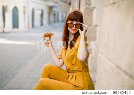 Street food, tourism, lunch time and vacation concept. Young smiling dark haired Caucasian girl in yellow suit and sunglasses, eating croissant while sitting on the stairs in the city. Copy space 69985463