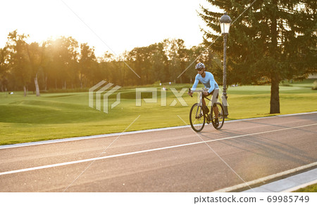 Athletic man in sportswear cycling in city park at sunset, riding bicycle along a road Athletic man in sportswear cycling in city park at sunset, riding bicycle along a road 69985749