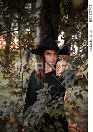 Young woman in dark dress and witch's hat holds lantern with candles in her hands and illuminates the forest. Halloween party costume. Park with autumn trees. Young woman in dark dress and witch's hat holds lantern with candles in her hands and illuminates the forest. Halloween party costume. Park with autumn trees. 69985761