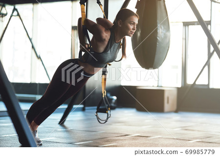 Young beautiful athletic woman wearing sports clothes exercising with fitness trx straps, doing push ups while training at gym Young beautiful athletic woman wearing sports clothes exercising with fitness trx straps, doing push ups while training at gym 69985779