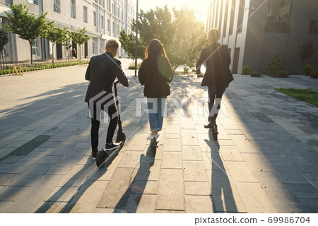 Back view of joyful international friends holding electric scooters going near modern building and talking. 69986704
