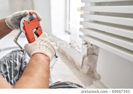 Plumber cuts the desired length of pipe with scissors tool. connection heating pipes to white Radiator in a new apartment under construction. Plumber cuts the desired length of pipe with scissors tool. connection heating pipes to white Radiator in a new apartment under construction. 69987393