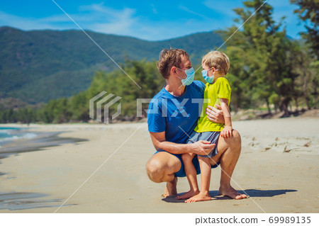 Normal reality during COVID. Father squatting and holding son looking on each other, wearing face blue mask protect coronavirus even walking nature sand sea beach near forest park sun day 69989135