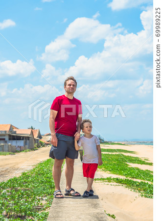 Father son walk along narrow concrete path, sea blue clear sky green grass background. Symbol as dad leads child into future adulthood. Happy childhood, fathers day, daddy influence on boy worldview Father son walk along narrow concrete path, sea blue clear sky green grass background. Symbol as dad leads child into future adulthood. Happy childhood, fathers day, daddy influence on boy worldview 69989225