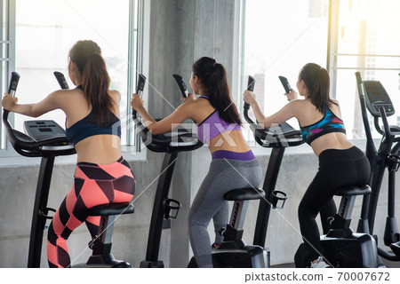 Rear View Of Young Women Sitting On Exercise Bikes In Health Club Rear View Of Young Women Sitting On Exercise Bikes In Health Club 70007672