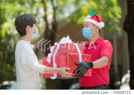 Asian delivery man wearing face mask and gloves and Christmas hat delivering present during COVID-19 Asian delivery man wearing face mask and gloves and Christmas hat delivering present during COVID-19 70009715