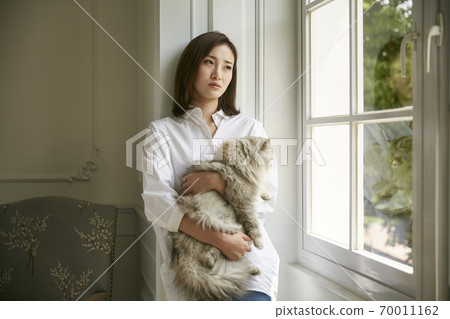 beautiful young asian woman standing by window at home holding cat in arms looking depressed and sad beautiful young asian woman standing by window at home holding cat in arms looking depressed and sad 70011162