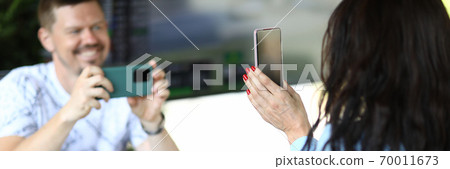 Woman is holding smartphone in her hands, man is filming review of novelty on smartphone in cafe. Woman is holding smartphone in her hands, man is filming review of novelty on smartphone in cafe. 70011673
