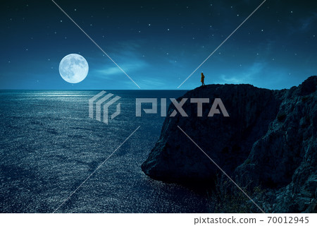 Person is standing in the edge of a cliff against the sea and full moon, under stars and moon light. Person is standing in the edge of a cliff against the sea and full moon, under stars and moon light. 70012945