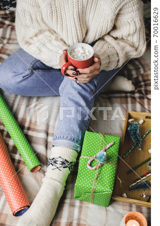 Young woman sits on plaid in cozy knitted woolen white sweater and holds cup of cocoa with marshmallows. Hygge New Year, preparation for holidays. Candles, Christmas balls, gifts, wrapping paper. Young woman sits on plaid in cozy knitted woolen white sweater and holds cup of cocoa with marshmallows. Hygge New Year, preparation for holidays. Candles, Christmas balls, gifts, wrapping paper. 70016629