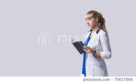 Confident young woman doctor standing with notebook or medical book in her hands. Cheks records, recipes or prescriptions. Intern in white medical clothes posing on light grey background. Confident young woman doctor standing with notebook or medical book in her hands. Cheks records, recipes or prescriptions. Intern in white medical clothes posing on light grey background. 70017006