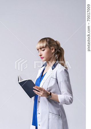 Confident young woman doctor standing with notebook or medical book in her hands. Cheks records, recipes or prescriptions. Intern in white medical clothes posing on light grey background. Confident young woman doctor standing with notebook or medical book in her hands. Cheks records, recipes or prescriptions. Intern in white medical clothes posing on light grey background. 70017009