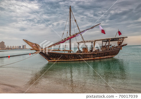 Traditional wooden boat dhow in Katara beach Qatar with Qatar flag and clouds in the sky in background 70017039
