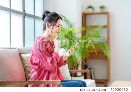 一個女人在沙發上放鬆的同時操作智能手機。 一個女人在沙發上放鬆的同時操作智能手機。 70030980