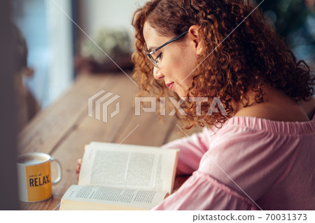 Young red hair woman with eyeglasses is sitting and reading a book Young red hair woman with eyeglasses is sitting and reading a book 70031373