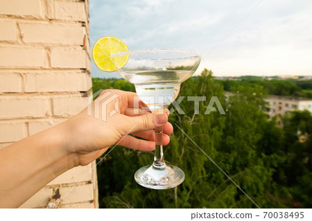 Stay at home balcony cocktail party. Classic home-made Margarita alcohol drink with lime and salt in a hand, trees sky and residential area houses view. Good for website or blog recipes or articles. Stay at home balcony cocktail party. Classic home-made Margarita alcohol drink with lime and salt in a hand, trees sky and residential area houses view. Good for website or blog recipes or articles. 70038495