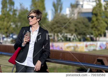 Portrait of a business woman leaning on railing with red bag and takeaway coffee in hands 70038560