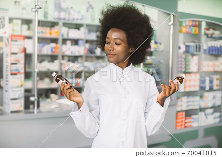 Which medicine is better, pharmacy, drug concept. Pretty African woman pharmacist or chemist choosing between two drugs in bottles, while standing in interior of modern pharmacy Which medicine is better, pharmacy, drug concept. Pretty African woman pharmacist or chemist choosing between two drugs in bottles, while standing in interior of modern pharmacy 70041015