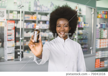 Confident smiling African woman, medical physician doctor or pharmacist posing over hospital pharmacy background, holding bottle of medicine in hands. Focus on face Confident smiling African woman, medical physician doctor or pharmacist posing over hospital pharmacy background, holding bottle of medicine in hands. Focus on face 70041016