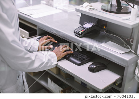 Cropped close up image of hands of African female clinical pharmacist, typing on computer keyboard, while standing behind the counter of modern hospital pharmacy Cropped close up image of hands of African female clinical pharmacist, typing on computer keyboard, while standing behind the counter of modern hospital pharmacy 70041033