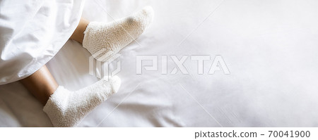 Woman feet in warm woolen socks on white bedclothes. Close up of young woman lying in bed at home. Woman feet in warm woolen socks on white bedclothes. Close up of young woman lying in bed at home. 70041900