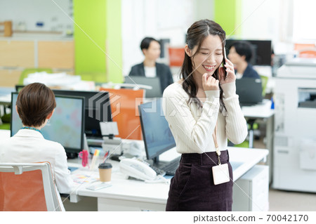 一名微笑的年輕女僱員談話在一個智能手機在辦公室 一名微笑的年輕女僱員談話在一個智能手機在辦公室 70042170