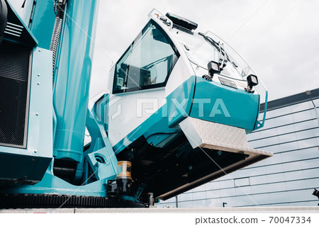 a cab with the operator of a Large blue car crane that stands ready to operate on hydraulic supports on a platform next to a large modern building. The largest truck crane for solving complex tasks. a cab with the operator of a Large blue car crane that stands ready to operate on hydraulic supports on a platform next to a large modern building. The largest truck crane for solving complex tasks. 70047334
