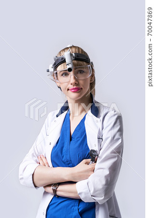 Confident ENT doctor wearing surgical headlight head light and protective glasses. Portrait of female otolaryngologist or head and neck surgeon standing arms crossed on light grey background. 70047869