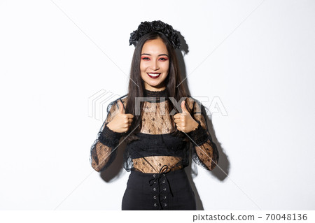 Image of happy asian woman in halloween party dress showing thumbs-up in approval. Girl wearing as witch or widow looking pleased, like or agree with something, white background 70048136
